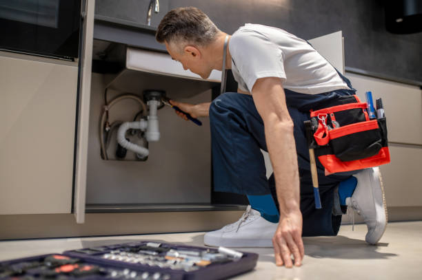 Eliminating problem. Middle aged man in t-shirt and overalls kneeling with wrench touching pipe under sink in lighted kitchen